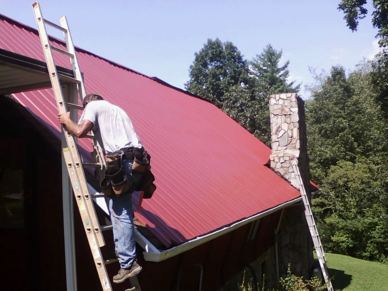 Roofer on a ladder beside a red metal roof and stone chimney — Soffit & Fascia Repair by Roofing Brothers in Allons